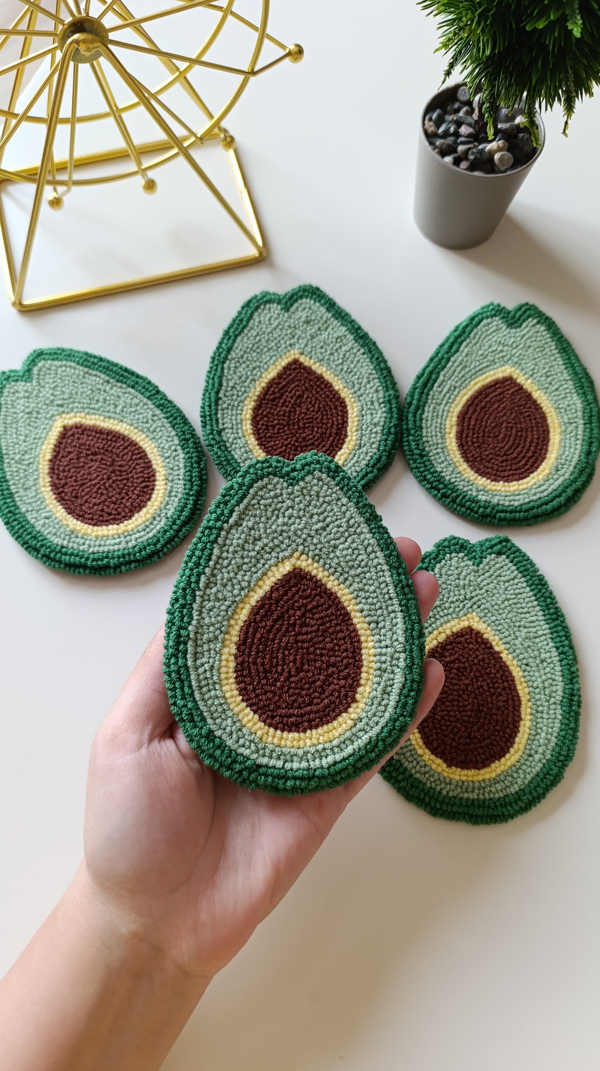 Five crocheted avocado half coasters arranged on a white surface, with a hand holding the front coaster. A gold wire decor object and a small potted plant are visible in the background.