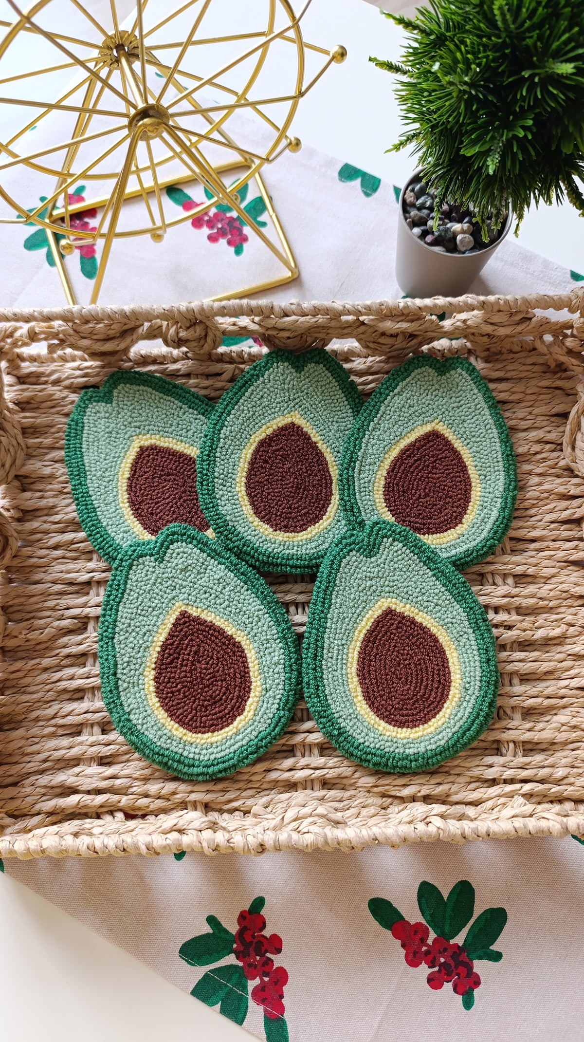 Four crocheted avocado slice coasters in green, beige, and brown, arranged on a woven straw tray with decorative props in the background.