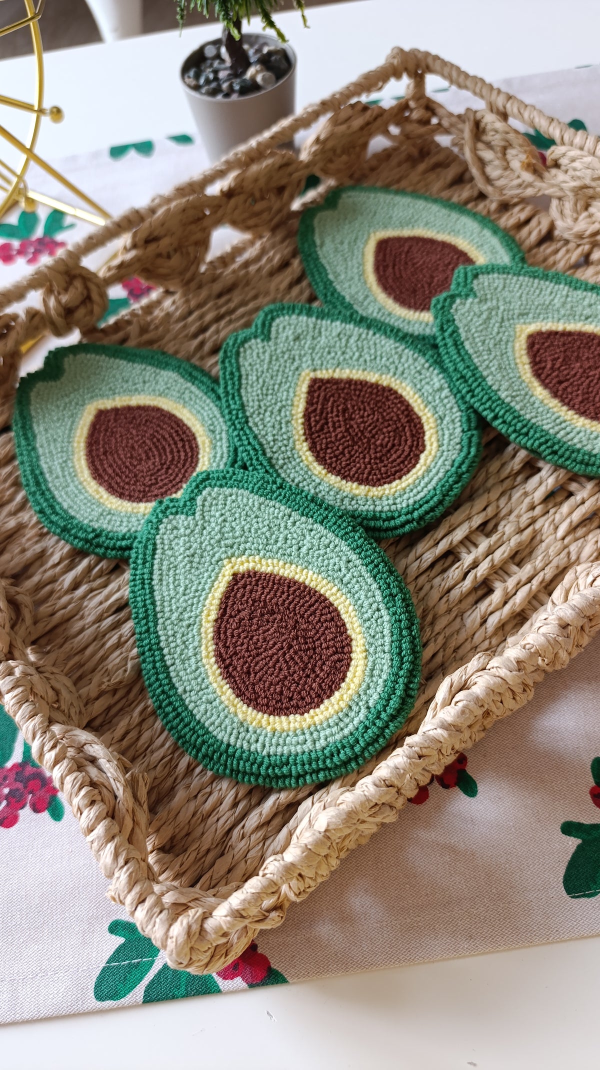 Four crocheted avocado half coasters in shades of green, cream, and brown, arranged on a woven straw tray over a floral tablecloth.