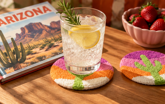 A clear textured drink coaster with a white, orange, and green cactus design holds a glass of iced water with a lemon slice and rosemary sprig, set on a wooden table next to an Arizona-themed book and a bowl of strawberries.