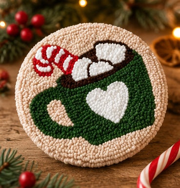A round punch needle craft piece depicting a festive hot cocoa scene, featuring a green mug with a white heart, a red-and-white striped candy cane, and white marshmallows in a dark brown cup, set on a wooden surface with holiday decorations.
