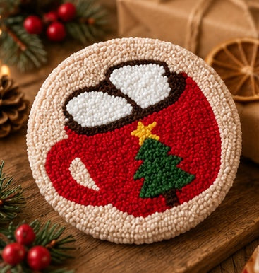 A round punch needle craft piece featuring a festive holiday design, including a red mug with white steam, a green Christmas tree, and a yellow star, displayed on a wooden surface with holiday decorations.