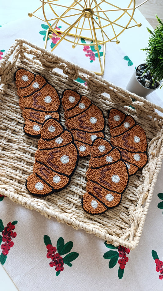 Three crocheted brown croissant coasters with white polka dots are arranged in a woven straw tray on a floral tablecloth.
