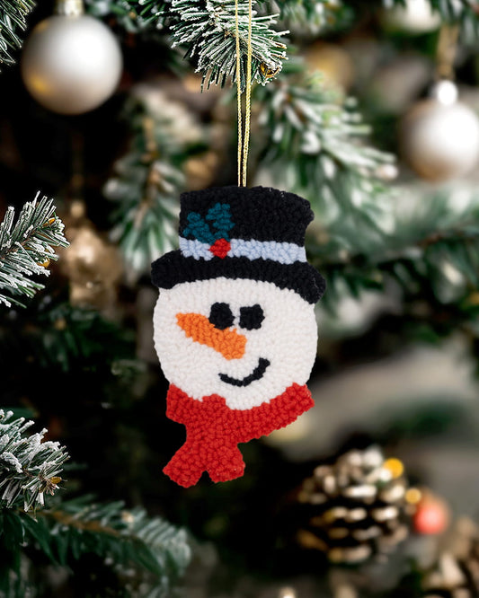 Handmade punch needle snowman ornament with black hat and red scarf hanging on a decorated Christmas tree.