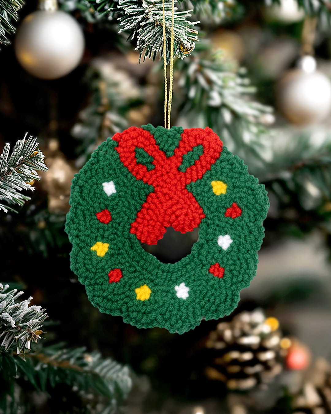 Handmade punch needle Christmas wreath ornament with red bow hanging on a decorated Christmas tree.