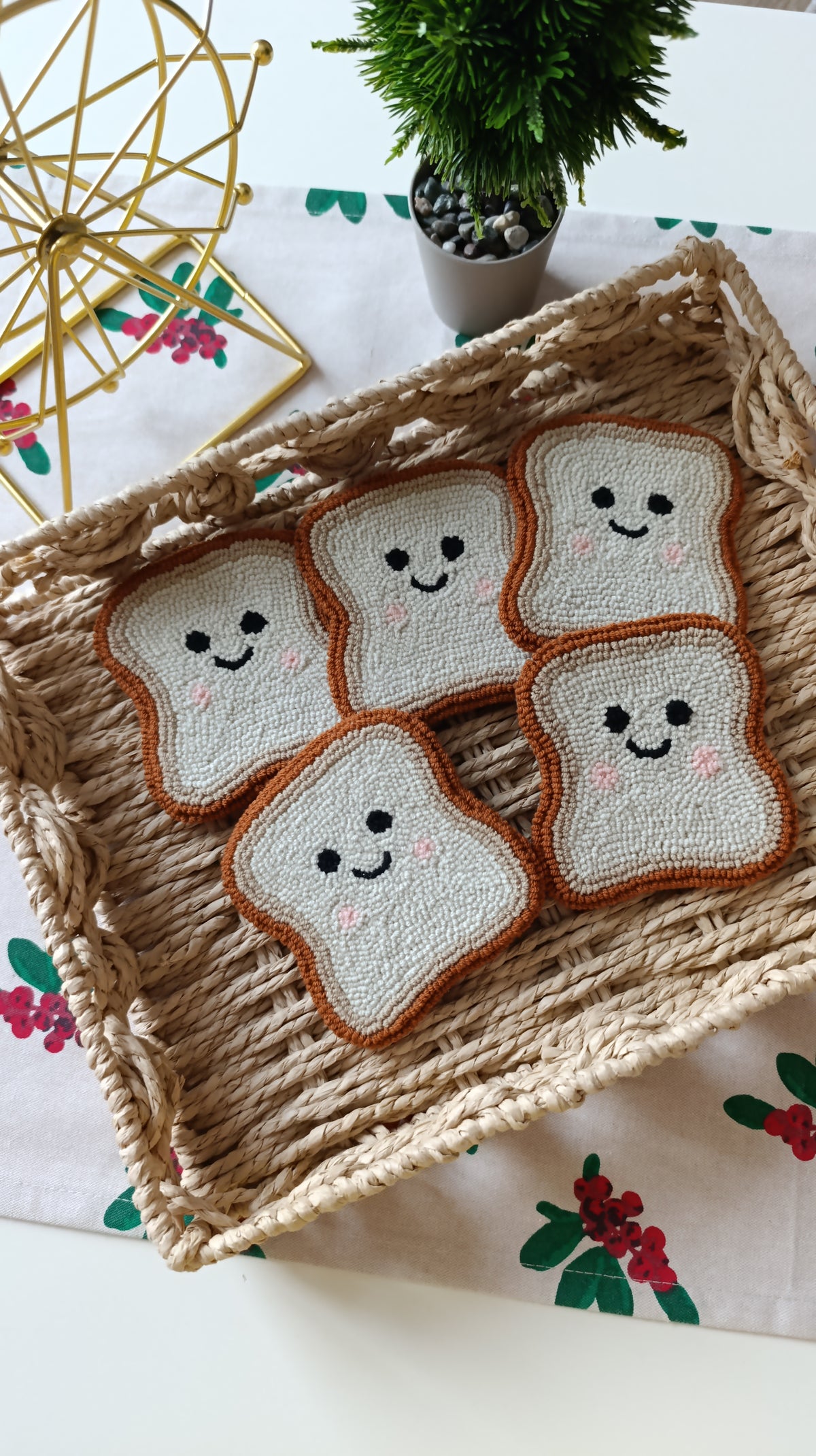 Five crocheted or knitted bread-slice shapes with smiling faces are arranged in a woven straw basket on a white table runner with a floral print. A small potted green plant and a gold geometric decor object are visible in the background.