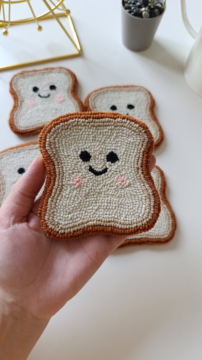 A hand holds a crocheted or tufted toast-shaped coaster in the foreground, with several matching toast coasters arranged on a white tabletop in the background. The coasters are cream/off-white with brown edges and simple embroidered or stitched smiling faces with pink cheeks.