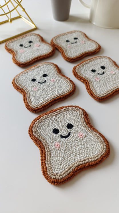 Five hand-punched toast-shaped coasters with embroidered smiling faces, arranged on a white tabletop with a gold geometric decor piece and mugs in the background.