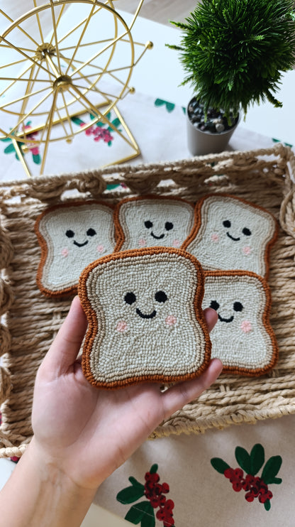 A hand holds a crocheted or knitted bread-slice-shaped patch with a smiling face, with several similar patches arranged on a woven tray in the background.