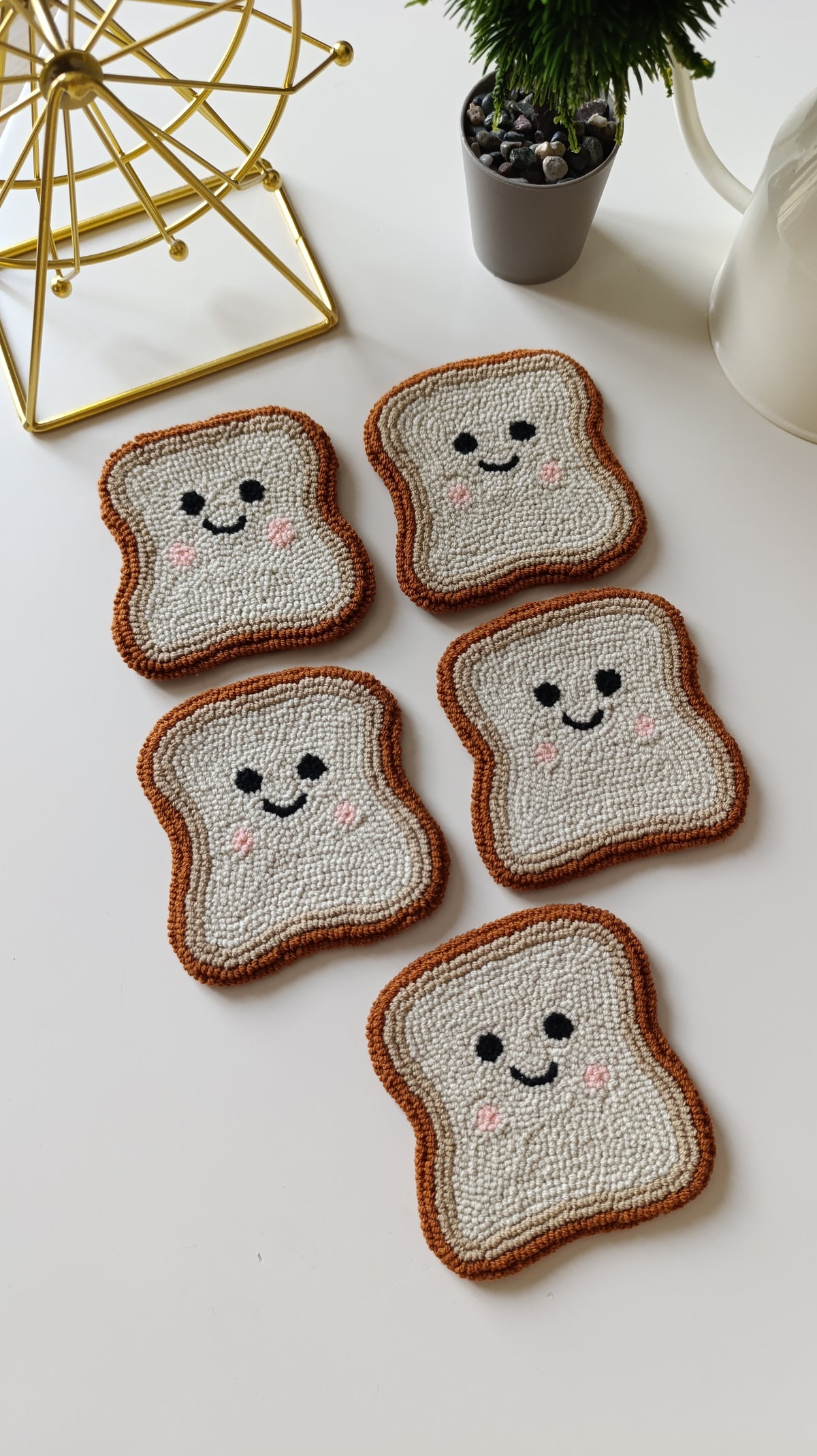 Five knitted toast-shaped coasters with smiling faces, arranged on a white tabletop with a small plant and decorative gold object in the background.